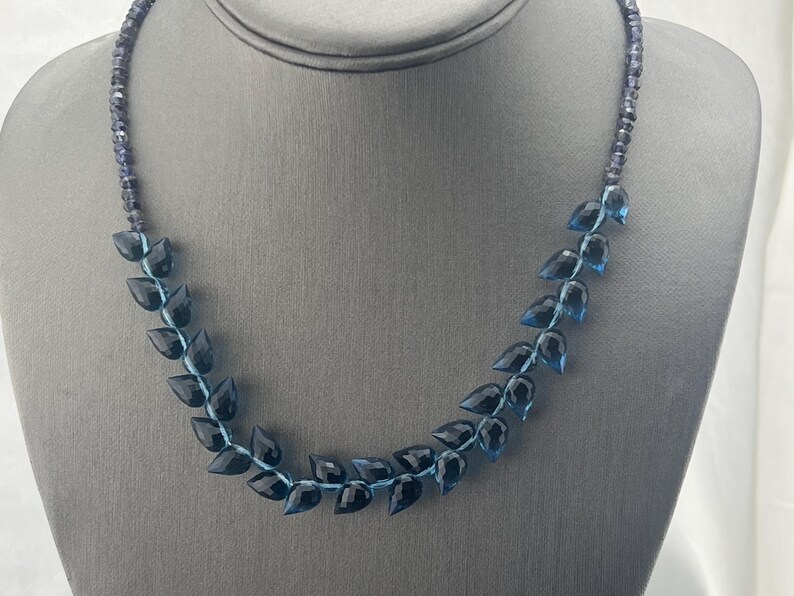 May include: A necklace featuring a leaf-like design. The necklace has dark blue, faceted gemstone leaves strung on a light blue cord. The necklace is displayed on a gray mannequin bust.