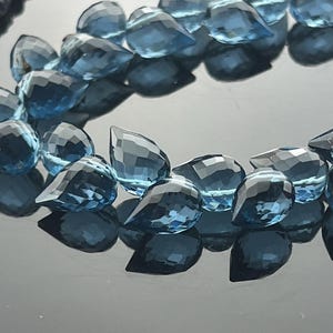 May include: A close-up of a strand of faceted, teardrop-shaped blue gemstones. The gems are arranged in a curved line, reflecting light and creating a sparkling effect. The background is a dark, reflective surface.