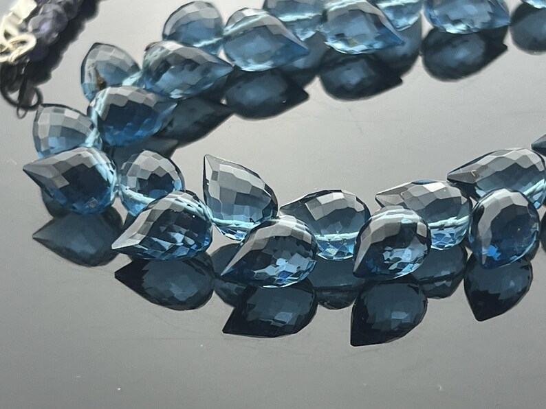 May include: A close-up of a strand of faceted, teardrop-shaped blue gemstones. The gems are arranged in a curved line, reflecting light and creating a sparkling effect. The background is a dark, reflective surface.