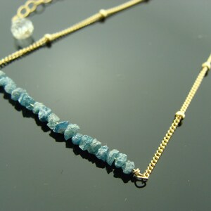 May include: A gold chain necklace with a row of light blue rough diamonds. The chain has small gold beads.