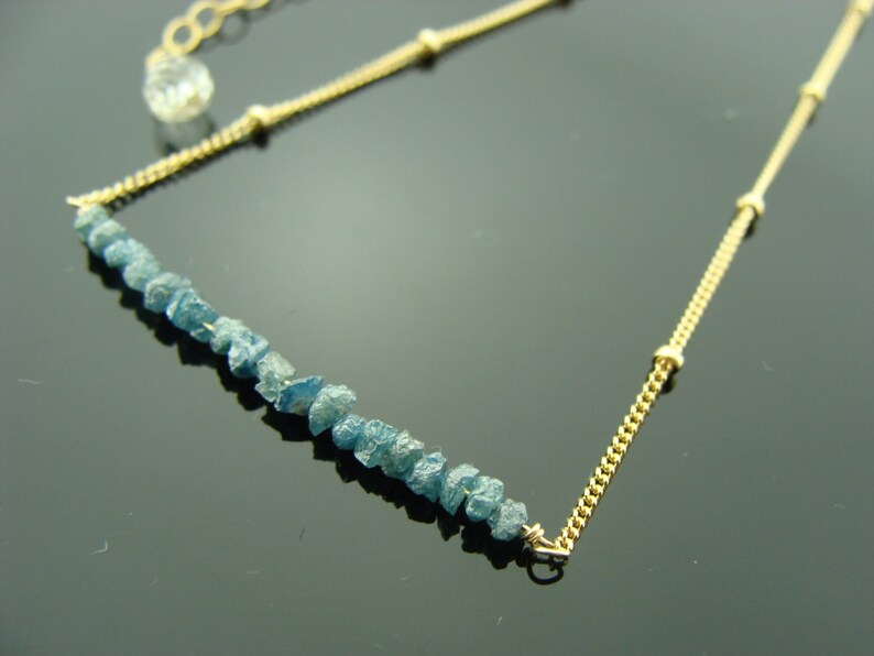 May include: A gold chain necklace with a row of light blue rough diamonds. The chain has small gold beads.