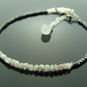 May include: A delicate bracelet featuring a row of rough, white diamonds and a row of black diamonds. The bracelet has a silver clasp and a small, white gemstone charm.