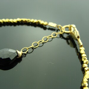May include: A gold bracelet with a black gemstone pendant and a gold clasp. The bracelet is made of small, gold beads.