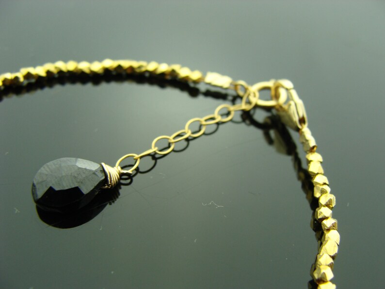 May include: A gold bracelet with a black gemstone pendant and a gold clasp. The bracelet is made of small, gold beads.