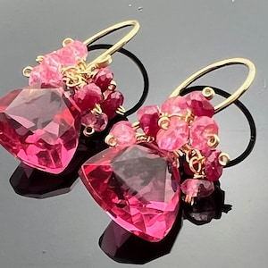 May include: A pair of dangle earrings featuring large, faceted pink gemstones. Smaller pink and red gemstones cluster around the larger stones, connected by gold-colored wire. The earrings have gold-colored ear wires and black accents.