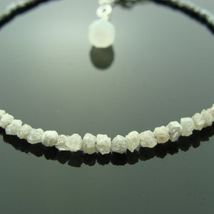 May include: A delicate necklace featuring a row of rough, white diamonds and a small, white gemstone pendant. The necklace is strung on a black cord.