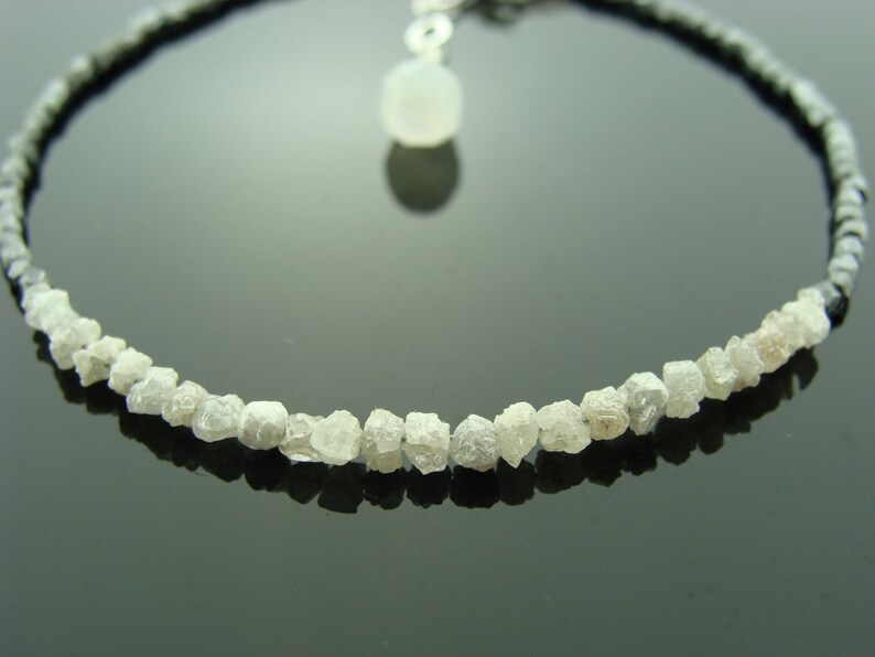 May include: A delicate necklace featuring a row of rough, white diamonds and a small, white gemstone pendant. The necklace is strung on a black cord.