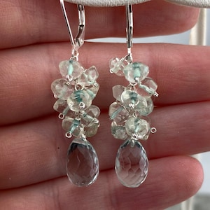 May include: Silver dangle earrings featuring a cluster of light blue and clear faceted gemstones. The earrings have a teardrop-shaped gemstone at the bottom.