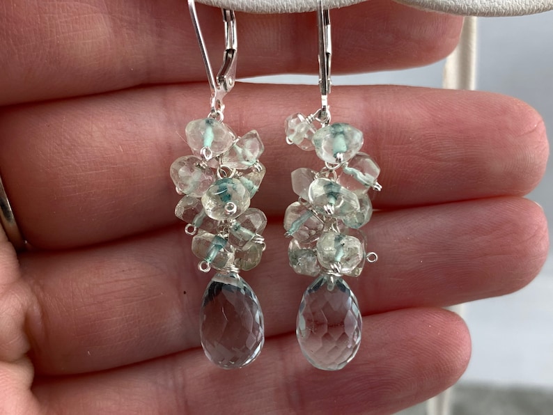 May include: Silver dangle earrings featuring a cluster of light blue and clear faceted gemstones. The earrings have a teardrop-shaped gemstone at the bottom.