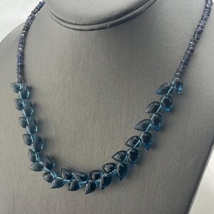 May include: A blue gemstone necklace displayed on a gray mannequin. The necklace features a strand of small, dark blue beads and a series of larger, teardrop-shaped blue gemstones. The gemstones graduate in color from light to dark.
