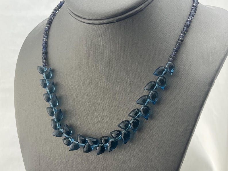 May include: A blue gemstone necklace displayed on a gray mannequin. The necklace features a strand of small, dark blue beads and a series of larger, teardrop-shaped blue gemstones. The gemstones graduate in color from light to dark.
