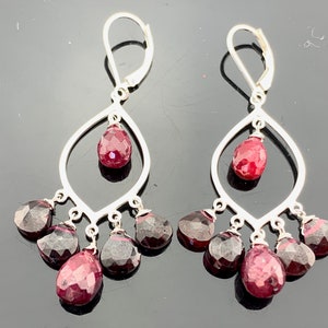 Puede incluir: Un par de pendientes de plata con gotas de piedras preciosas rojas y rojo oscuro. Los pendientes tienen un marco en forma de lágrima con múltiples gotas más pequeñas que cuelgan de la parte inferior.