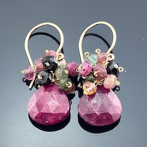 May include: A pair of gold-colored drop earrings. Each earring features a cluster of small, multi-colored beads above a large, faceted, teardrop-shaped, magenta-colored stone. The earrings have a simple hook design.