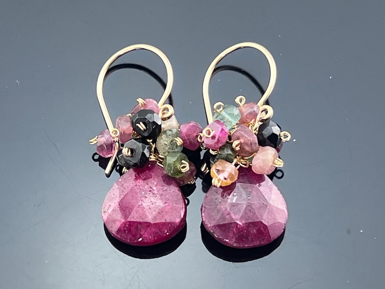 May include: A pair of gold-colored drop earrings. Each earring features a cluster of small, multi-colored beads above a large, faceted, teardrop-shaped, magenta-colored stone. The earrings have a simple hook design.