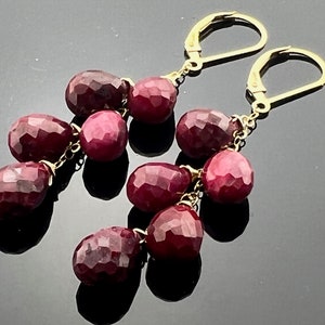 May include: A pair of gold earrings with dangling ruby gemstones. The earrings are teardrop shaped and connected by a delicate chain.