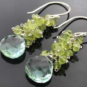Op de afbeelding: Zilveren oorhangers met een traanvormige lichtgroene edelsteen en een cluster van kleinere lichtgroene edelstenen.