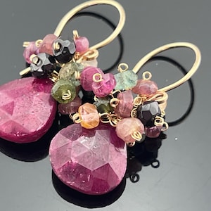 May include: A pair of gold-colored dangle earrings. Each earring features a large, faceted, teardrop-shaped, deep pink gemstone. Clusters of smaller, multi-colored gemstones in pink, green, and black are attached above the teardrops. The earrings have gold-colored ear wires.