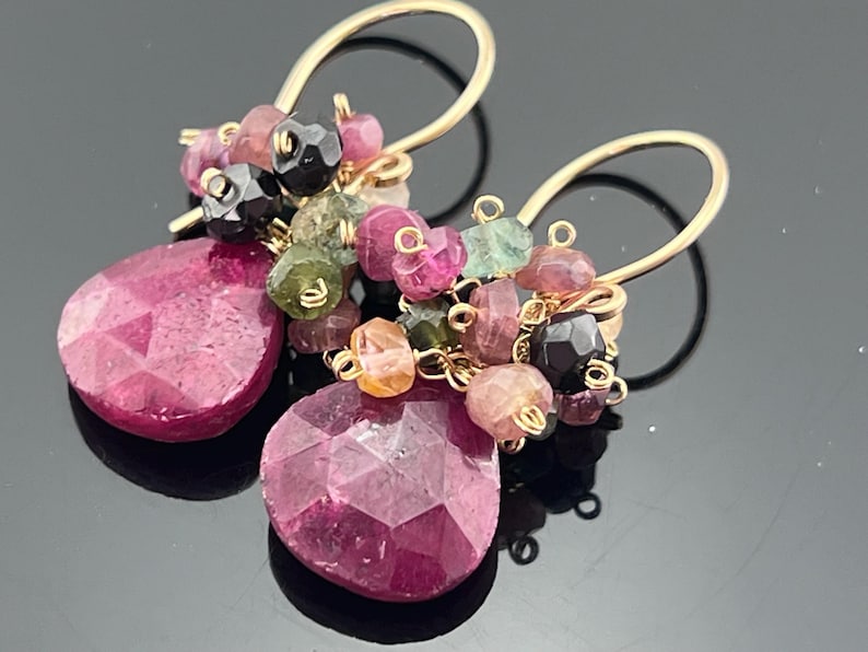 May include: A pair of gold-colored dangle earrings. Each earring features a large, faceted, teardrop-shaped, deep pink gemstone. Clusters of smaller, multi-colored gemstones in pink, green, and black are attached above the teardrops. The earrings have gold-colored ear wires.