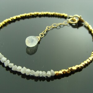 May include: A gold bracelet with a row of small, rough diamonds and a row of gold beads. A single, teardrop-shaped, clear gemstone hangs from the bracelet.