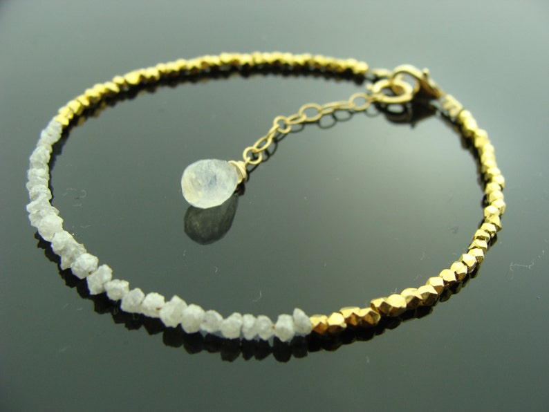 May include: A gold bracelet with a row of small, rough diamonds and a row of gold beads. A single, teardrop-shaped, clear gemstone hangs from the bracelet.