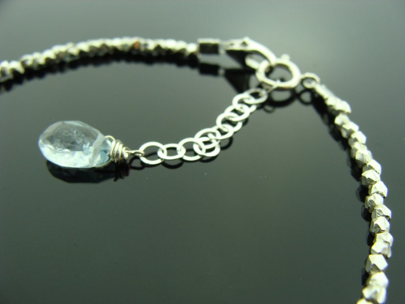 May include: A silver bracelet with a light blue gemstone pendant. The bracelet is made of small, faceted beads and has a lobster claw clasp.