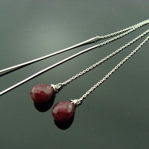 May include: A pair of silver dangle earrings with red gemstone drops. The earrings have a long, thin chain that hangs from a small silver loop.