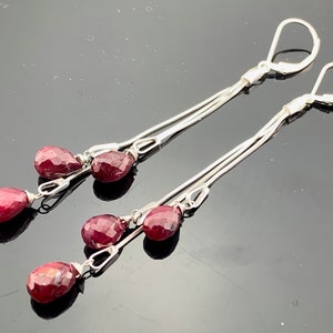 May include: A pair of dangle earrings featuring faceted, teardrop-shaped red gemstones. The earrings have a silver-toned metal design with a lever-back closure. The gemstones are suspended from delicate chains, creating a graceful, elongated silhouette.