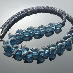 May include: A two-strand necklace featuring dark blue beads and faceted blue teardrop-shaped gemstones. The necklace has a silver clasp and is displayed on a reflective surface.