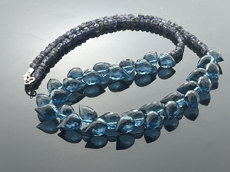 May include: A two-strand necklace featuring dark blue beads and faceted blue teardrop-shaped gemstones. The necklace has a silver clasp and is displayed on a reflective surface.