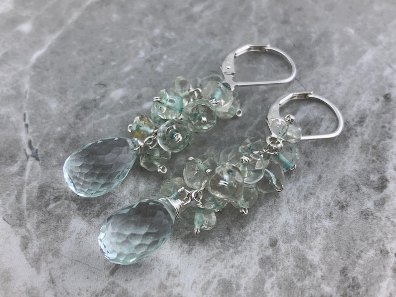 May include: A pair of silver dangle earrings with a teardrop-shaped aquamarine gemstone at the bottom of each earring. The earrings are made with a cluster of smaller aquamarine gemstones.