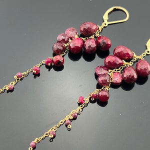 May include: A pair of gold earrings with a cluster of ruby red gemstones dangling from each earring. The earrings have a delicate chain with smaller ruby red gemstones hanging from the chain.