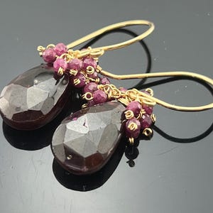 May include: Gold dangle earrings with a teardrop-shaped dark purple gemstone and a cluster of smaller pink gemstones.