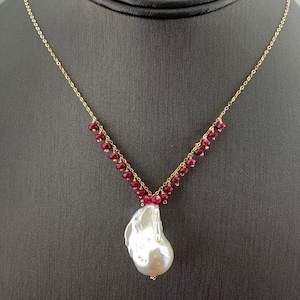May include: A gold chain necklace with a large white pearl pendant and a V-shaped design of small red beads.