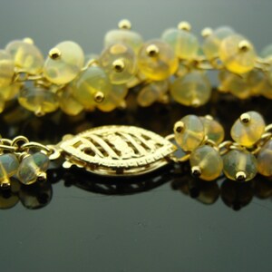 May include: Close-up of a gold necklace with multiple strands of small, translucent, yellow-green beads. The necklace features a gold clasp with a leaf-like design. The beads are clustered together, creating a delicate and elegant appearance.