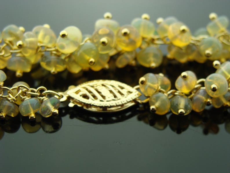 May include: Close-up of a gold necklace with multiple strands of small, translucent, yellow-green beads. The necklace features a gold clasp with a leaf-like design. The beads are clustered together, creating a delicate and elegant appearance.