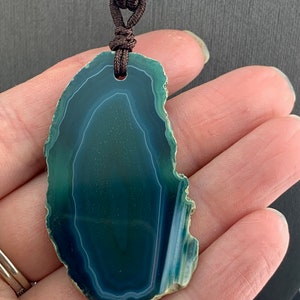 Pu&ograve; includere: Un ciondolo in agata verde con una collana in corda marrone. L'agata ha una forma naturale e organica e presenta un motivo a spirale di verde e bianco.