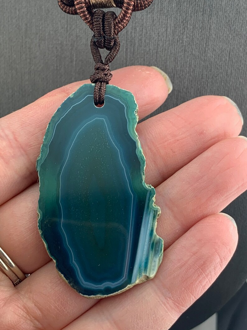 Pu&ograve; includere: Un ciondolo in agata verde con una collana in corda marrone. L'agata ha una forma naturale e organica e presenta un motivo a spirale di verde e bianco.