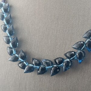 May include: A necklace featuring faceted, teardrop-shaped beads in shades of blue and black. The beads are strung together with a light blue cord, creating a delicate and elegant jewelry piece. The necklace is displayed against a neutral gray background.