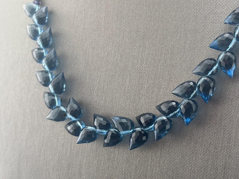 May include: A necklace featuring faceted, teardrop-shaped beads in shades of blue and black. The beads are strung together with a light blue cord, creating a delicate and elegant jewelry piece. The necklace is displayed against a neutral gray background.