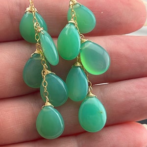 May include: Green gemstone dangle earrings with gold chain and wire accents. The earrings feature multiple teardrop-shaped gemstones.