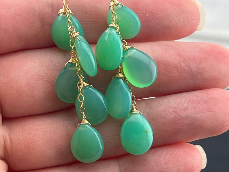 May include: Green gemstone dangle earrings with gold chain and wire accents. The earrings feature multiple teardrop-shaped gemstones.