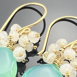 May include: Gold dangle earrings featuring a light blue gemstone and white stones. The earrings are crafted with wire wrapped stones.