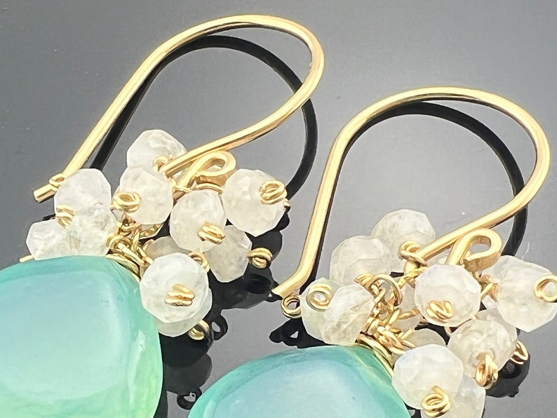 May include: Gold dangle earrings featuring a light blue gemstone and white stones. The earrings are crafted with wire wrapped stones.