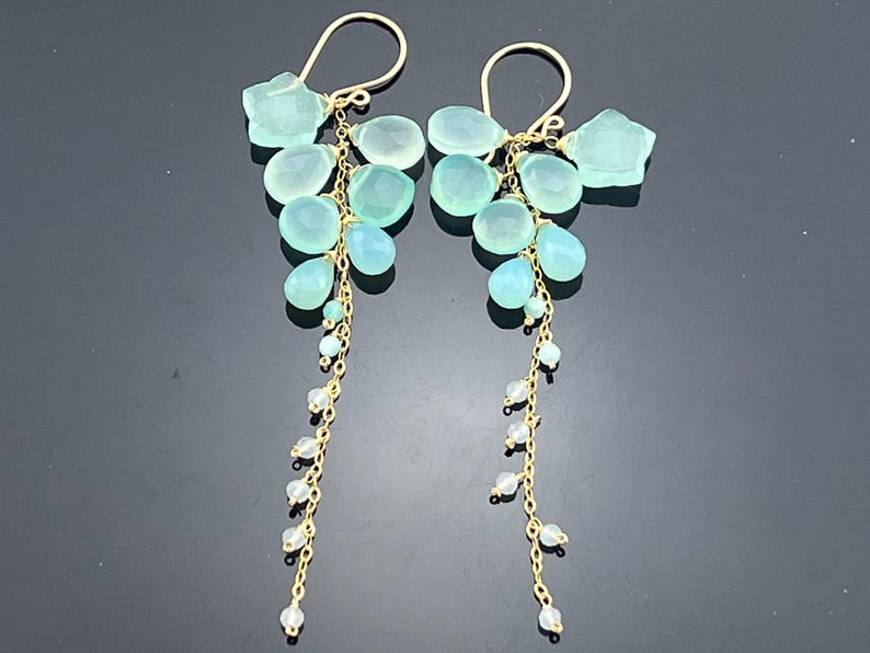 May include: A pair of elegant dangle earrings featuring a cluster of light blue gemstones and small white beads suspended from gold-colored hooks. The earrings have a delicate, cascading design, perfect for adding a touch of sophistication.