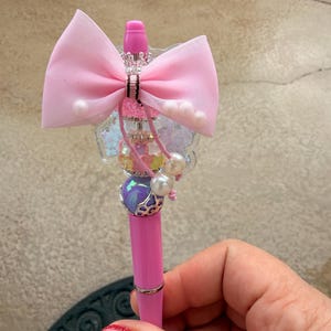 Puede incluir: Bolígrafo rosa con lazo decorativo, perlas y detalles iridiscentes. El bolígrafo presenta un lazo rosa, una corona plateada y una cuenta morada y plateada. El bolígrafo se sostiene en una mano con esmalte de uñas rojo brillante.