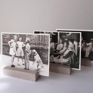 Könnte beinhalten: Ein Satz von vier Schwarz-Weiß-Fotografien, die auf rechteckigen Betonhaltern ausgestellt sind. Die Fotos zeigen Szenen von Kindern, einem Hund und Menschen in einem Café. Die Halter sind neutralgrau.