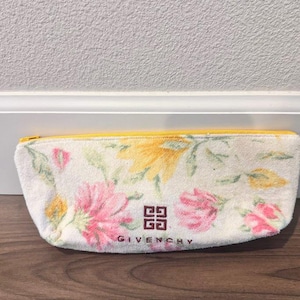 May include: A white cosmetic bag with a floral pattern and a yellow zip. The bag displays pink and yellow flowers and the Givenchy logo in brown. The rectangular bag appears to be made of a soft, absorbent material.