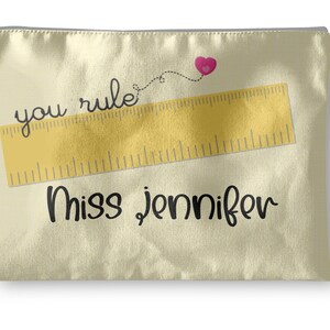 May include: Cream-colored zippered pouch with a gold ruler graphic and the words "you rule" and "Miss Jennifer" in black script. A pink heart is connected to the word "rule" with a dotted line.