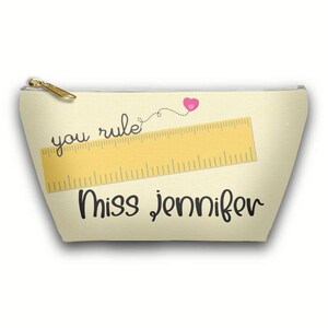 May include: A beige cosmetic bag with a gold zipper. The bag features a yellow ruler graphic, the words "you rule" and "Miss Jennifer", and a pink heart.
