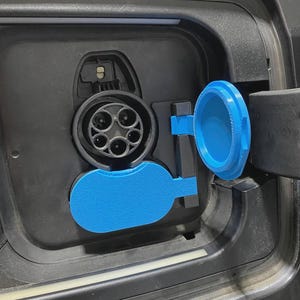 May include: Close-up of an electric vehicle charging port. The port is black with a circular charging socket and a blue protective cover. The cover is open, revealing the charging port.